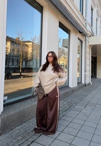 Oversize beige striktrøje, brune satin bukser med vide ben og hvide striber, grå skuldertaske. Reflekterende glasbygning i baggrunden.