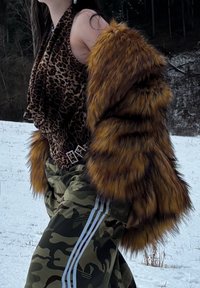 Femme appuyée en arrière à l'extérieur, portant un haut à imprimé léopard, un manteau en fausse fourrure marron rayé, et un pantalon camouflage avec des bandes blanches dans un décor enneigé.