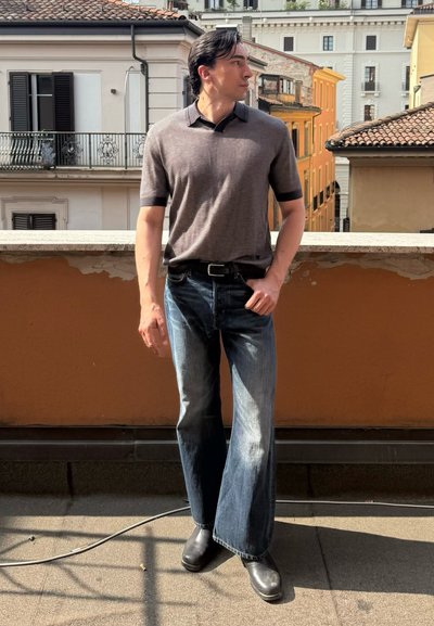 Camisa polo de manga corta en gris oscuro con detalles en negro, combinada con jeans anchos descoloridos de color azul y botas negras, de pie contra un fondo de balcón.