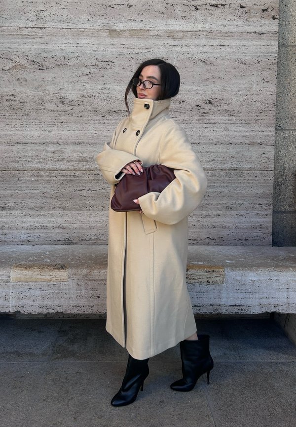 Frau in langem beigem Mantel mit hohem Kragen, schwarzen Stiefeln mit Absatz, Brille und einer braunen Clutch in der Hand, steht vor einer Steintapete.