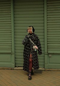 Svart og rosa mønstret kunstpelsjakke, kne-lengde, båret over en svart topp og strømpebukser. Rosa og svarte platåstøvler, bærende på kosedyr.