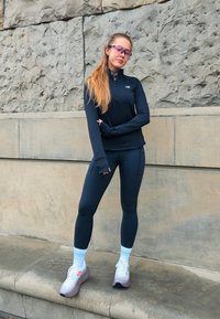 Noor naine mustades spordiriietes ja valgetes tossudes seisab kiviseina vastu, käed risti, kandes toonitud spordiprille.