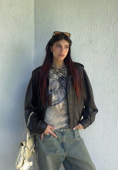 Joven con el pelo largo y oscuro, lleva gafas de sol en la cabeza, chaqueta de cuero negra, camiseta con diseño y vaqueros azules, sosteniendo un bolso de color claro.