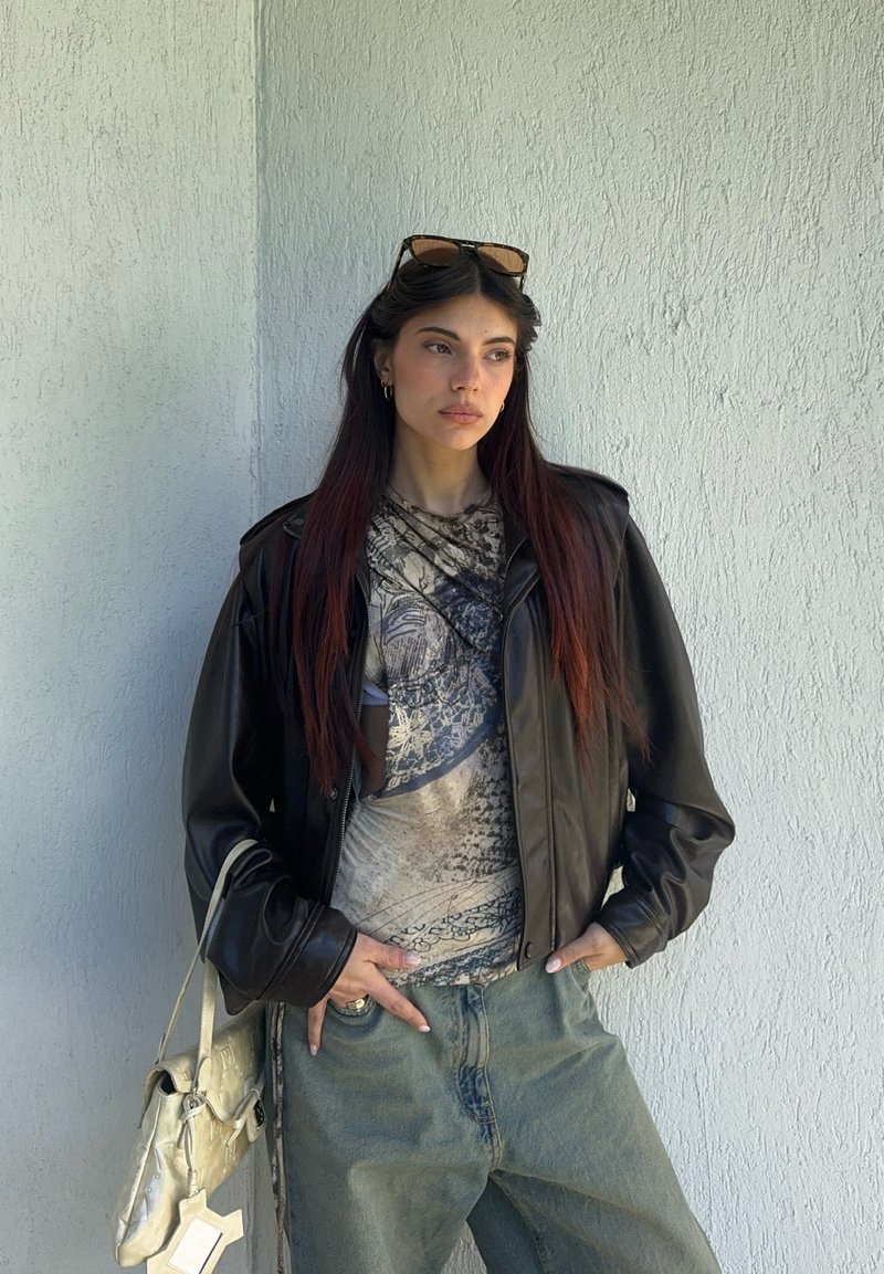 Giovane donna con capelli lunghi e scuri che indossa occhiali da sole sulla testa, giacca di pelle nera, maglietta con grafica e jeans blu, che tiene una borsa chiara.