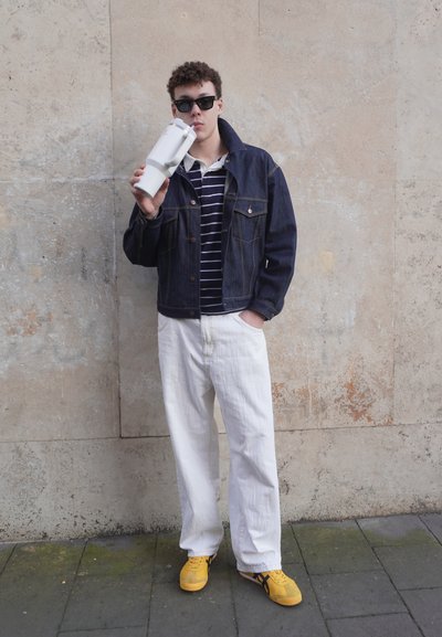 Chaqueta de mezclilla sobre una camisa a rayas, pantalones anchos blancos y zapatillas amarillas. Sosteniendo un recipiente blanco. Fondo texturizado en tonos neutros.