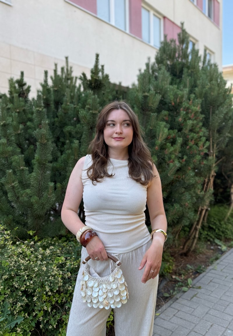 Jeune femme aux longs cheveux bruns tient un petit sac coquillage blanc, porte un top blanc sans manches et un pantalon, debout près des buissons verts en extérieur.