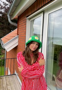 Cappello a secchiello verde all'uncinetto con accenti di margherita bianca, abbinato a una camicia a righe rossa e bianca con bottoni, posato davanti a un muro di mattoni.