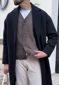 Mann trägt einen schwarzen Mantel, eine braune Strickjacke mit Knöpfen, ein weißes Hemd und beige Hosen, hält den Mantel offen und trägt eine weiße Tragetasche im Freien.