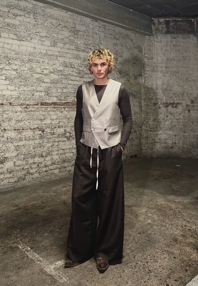 Persona con cabello rizado rubio, vistiendo un chaleco beige, camiseta de manga larga negra y pantalones anchos negros, de pie en una habitación con paredes de ladrillo y concreto texturizado.