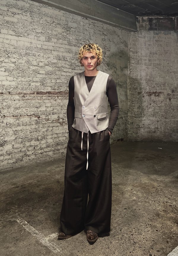 Άτομο με ξανθά, σγουρά μαλλιά, που φοράει μπεζ γιλέκο, μαύρη μπλούζα μακρυμάνικη και φαρδιά μαύρη παντελόνα, στέκεται σε ένα δωμάτιο με υφή από τούβλα και σκυρόδεμα.