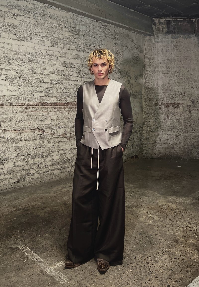 Personne aux cheveux blonds bouclés portant un gilet beige, un haut à manches longues noir et un pantalon large noir, debout dans une pièce en briques et béton texturés.