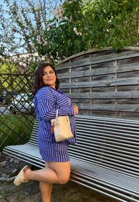 Vestito a camicia a righe blu e bianche, di lunghezza al ginocchio, con una tasca. Tiene in mano una piccola borsa color crema con un ciondolo. Siede su una panchina metallizzata.