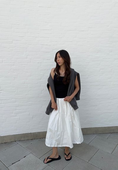 Top negro de tirantes, falda blanca con pliegues, sudadera gris sobre los hombros, chanclas negras. Piel clara, cabello castaño oscuro.