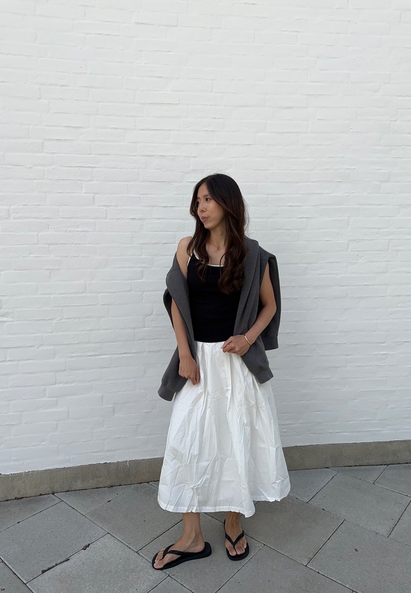 Μαύρο τοπ με τιράντες, λευκή πλισέ φούστα, γκρι φούτερ ριγμένο στους ώμους, μαύρες σαγιονάρες. Ανοιχτόχρωμη επιδερμίδα, σκούρα καστανά μαλλιά.