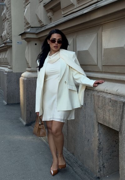 Mujer con vestido color marfil, blazer a juego sobre los hombros, tacones y bolso marrones, de pie contra una pared de piedra de un edificio.