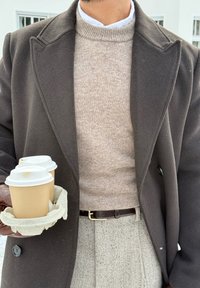 Homme en manteau marron, pull beige et pantalon à motifs tenant un plateau avec deux gobelets de café à emporter, debout en plein air.
