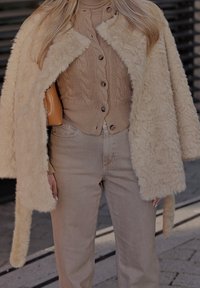 Γυναίκα που φοράει μπεζ πλεκτή ζακέτα και ζιβάγκο με παντελόνι ψηλόμεσο σε ταμπά, ντυμένη με μια αφράτη κρεμ παλτό, κρατώντας μια τσάντα σε ταμπά χρώμα.