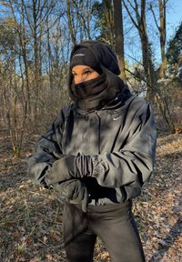 Giacca nera isolante con polsini elasticizzati, fascia multiplo al collo e guanti. Texture liscia con finitura opaca; ambiente esterno con alberi.