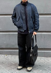 Άνδρας που φοράει σκούρο μπλε bomber μπουφάν, μαύρο παντελόνι με ευρεία πόδια, μπεζ παπούτσια και κρατάει μια μαύρη τσάντα ώμου, στέκεται μπροστά σε έναν γκρι τοίχο.