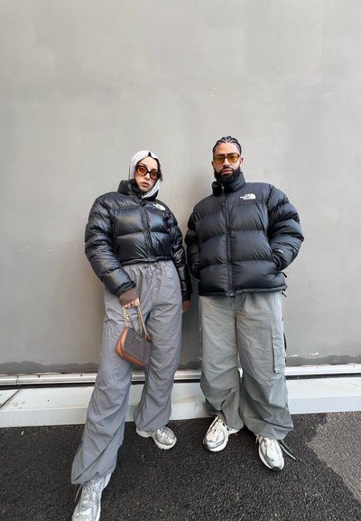 Dos individuos están de pie uno al lado del otro, vistiendo chaquetas de plumas negras, pantalones cargo grises y zapatillas deportivas. La mujer sostiene un bolso pequeño.