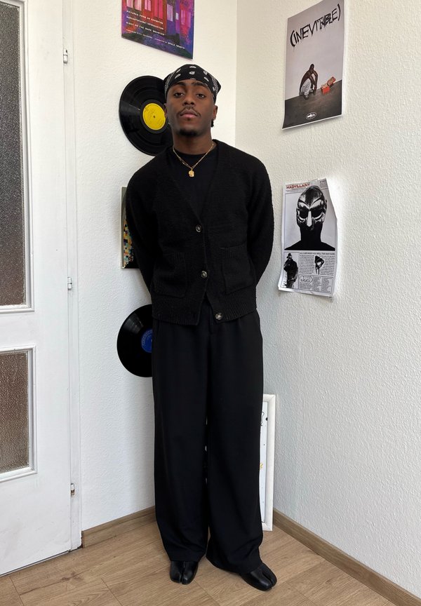 Črn kardigan z gumbi, črne dolge hlače in črne čevlje. Bandana na glavi, stoji ob steni z vinilnimi ploščami in posterji.