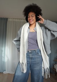 Szara oversizeowa bluza, lawendowy top na ramiączkach, szerokie niebieskie dżinsy i długi biały szal. Czarne nauszniki i złoty naszyjnik z zawieszką.