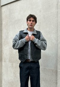 Sort lædervest med en grå fleecekrave, båret over en hvid skjorte, parret med mørke denimjeans. Modellen justerer vesten.