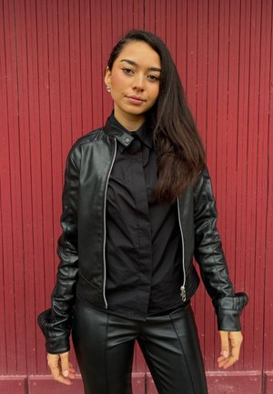 Joven con cabello largo y oscuro, vestida con chaqueta y pantalones de cuero negro, posando frente a un fondo rojo con paneles verticales.