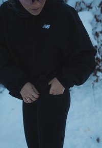 Black pullover jacket with a hood and elastic cuffs, paired with high-waisted black leggings. Hands placed in jacket pocket. Snowy background.