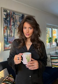 Jeune femme aux longs cheveux bruns, portant un haut en satin noir, tenant une tasse rose avec un motif de cactus, debout dans un salon confortable avec des coussins colorés.