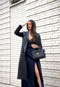 Femme en manteau en cuir noir et robe bleu marine fendue à la cuisse, tenant un sac à main noir, debout contre un mur en briques blanches.