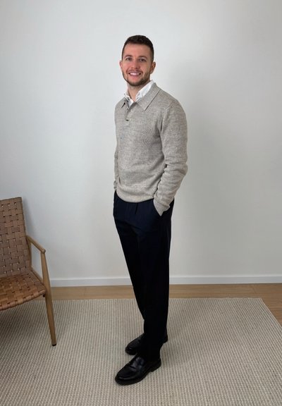 Hombre sonriente de pie con las manos en los bolsillos, vistiendo un suéter gris sobre una camisa blanca, pantalones negros y zapatos negros, junto a una pared blanca lisa y al lado de una silla marrón.