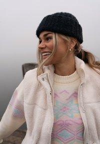 Veste en polaire beige clair portée sur un pull à motifs pastel, avec un bonnet tricoté foncé. Cheveux blonds lâches et boucles d'oreilles créoles dorées.