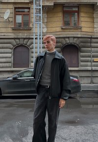 Sort læderjakke over grå rullekrave sweater, kombineret med mørke cowboybukser. Glatte teksturer; står på en våd gade nær en parkeret bil.