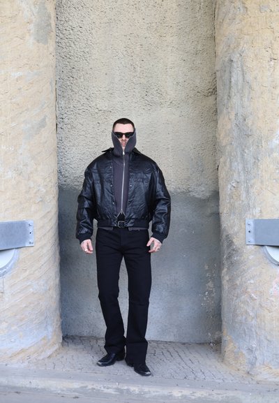 Hombre con chaqueta de cuero negro con cuello alto con cremallera, gafas de sol, pantalones negros y botas, de pie entre paredes beige texturizadas.