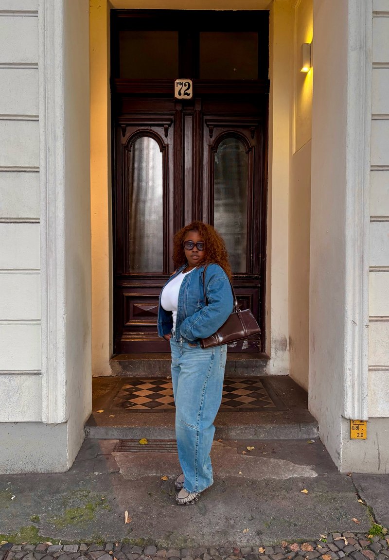 Casaco de ganga, t-shirt branca e jeans azul claro. Bolsa castanha e sapatos cinzentos. Portas de madeira escura com painéis de vidro ao fundo.