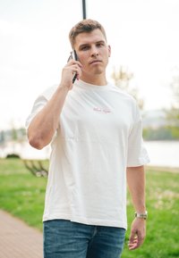 Vit t-shirt med kort ärm och texten "Black Roses" på bröstet, kombinerad med blå denimjeans. Personen håller en telefon.