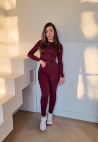 Burgundy fitted top and leggings made from stretchy material, paired with white trainers, posing next to a geometric white structure.