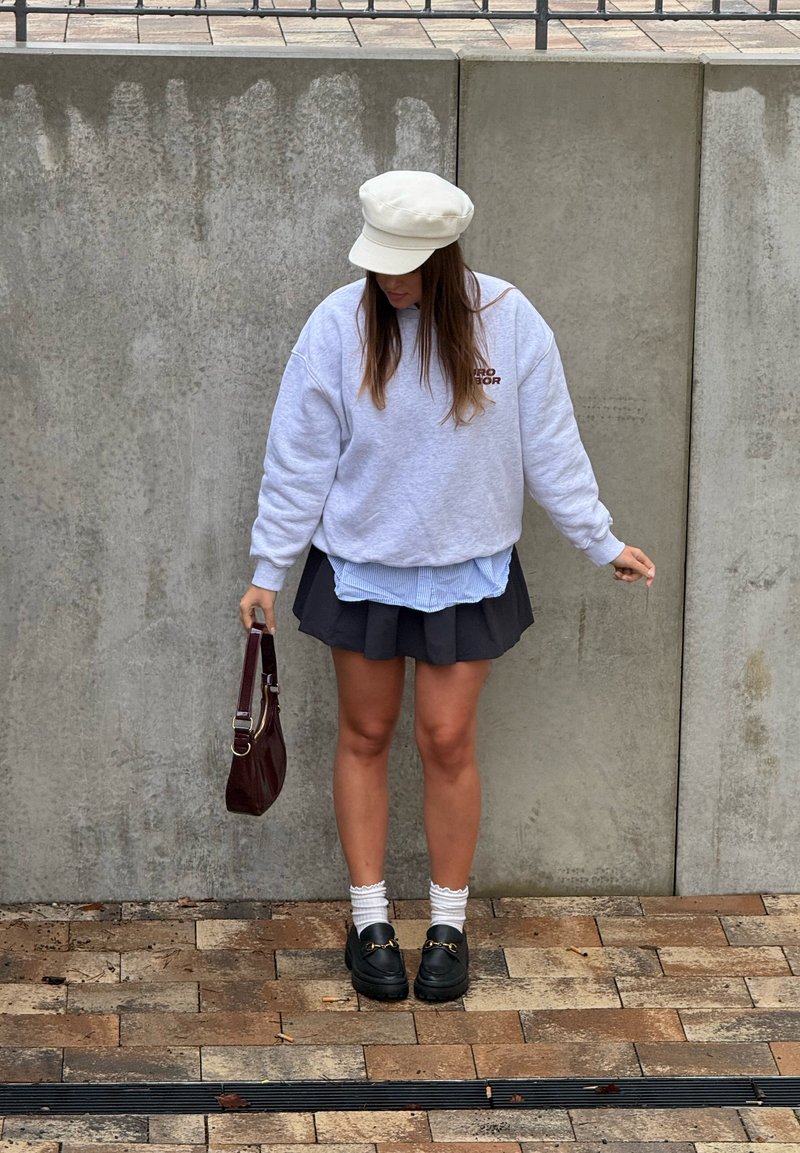 Femme portant une casquette blanche, un sweat-shirt gris, une jupe noire, des chaussettes blanches, des mocassins noirs, tenant un sac à main sombre contre un mur en béton.