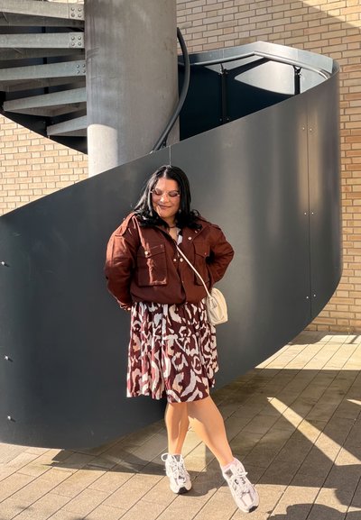 Mujer de pelo oscuro con gafas de sol, chaqueta marrón, vestido estampado y zapatillas blancas, de pie frente a una escalera de caracol negra al aire libre.