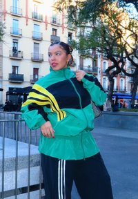 Femme portant un coupe-vent vert et noir avec des rayures jaunes, debout en plein air sur une place de la ville avec des arbres et des bâtiments en arrière-plan.