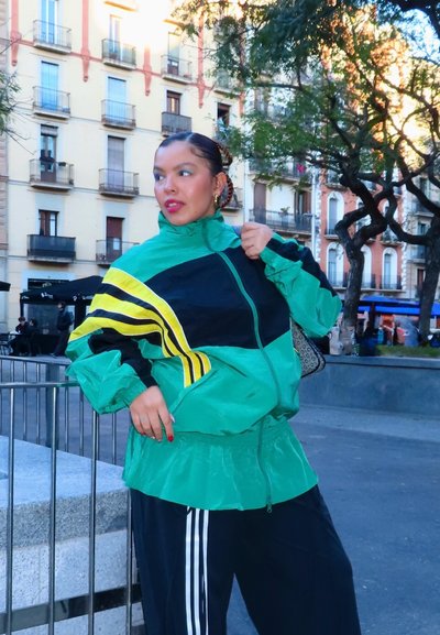 Mujer con cortavientos verde y negro con rayas amarillas, de pie al aire libre en una plaza de la ciudad con árboles y edificios al fondo.