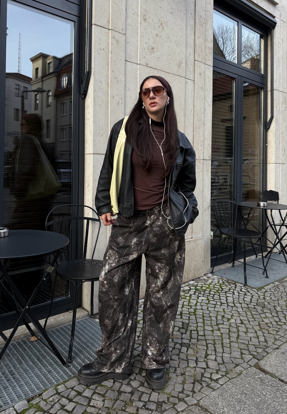 Veste en cuir noire sur un haut marron, pantalon large à motifs gris et noir, chaussures noires épaisses et lunettes de soleil ; debout près d'un café.