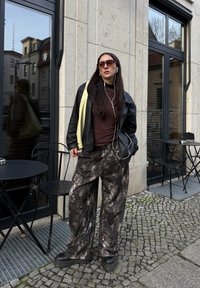 Veste en cuir noire sur un haut marron, pantalon large à motifs gris et noir, chaussures noires épaisses et lunettes de soleil ; debout près d'un café.