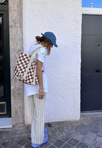 Canvas taske med røde og sorte designs på en cremefarvet baggrund, båret af en person i en hvid skjorte og stribede bukser.