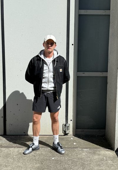 Chaqueta de mezclilla negra sobre una sudadera gris, pantalones cortos negros y zapatillas grises con calcetines blancos. El fondo presenta una pared gris clara.