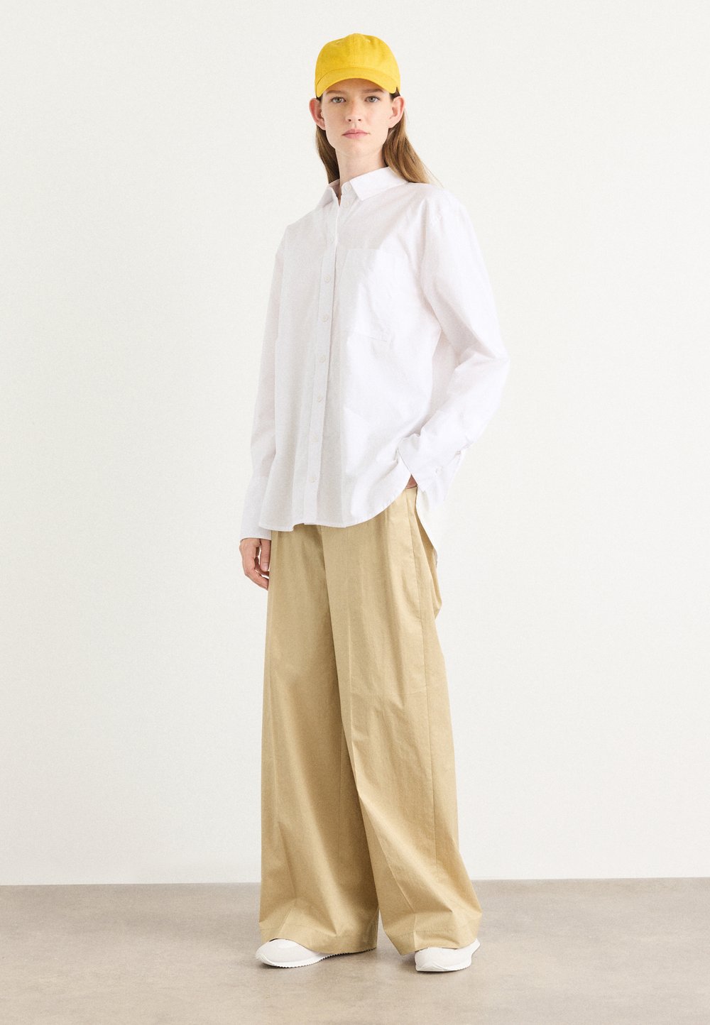 Camisa branca de botões com um bolso, combinada com calças largas de tom bege claro e sapatos brancos. Um boné amarelo completa o visual neutro.