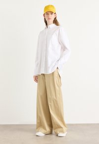 Camisa branca de botões com um bolso, combinada com calças largas de tom bege claro e sapatos brancos. Um boné amarelo completa o visual neutro.