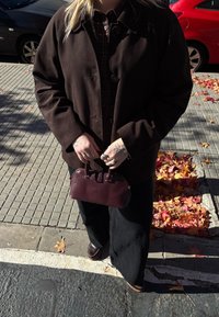 Hnedá semišová bunda so zapínaním na gombíky, pod ňou károvaná košeľa, čierne široké nohavice a malá bordová kabelka. Textúrované lístie na zemi.