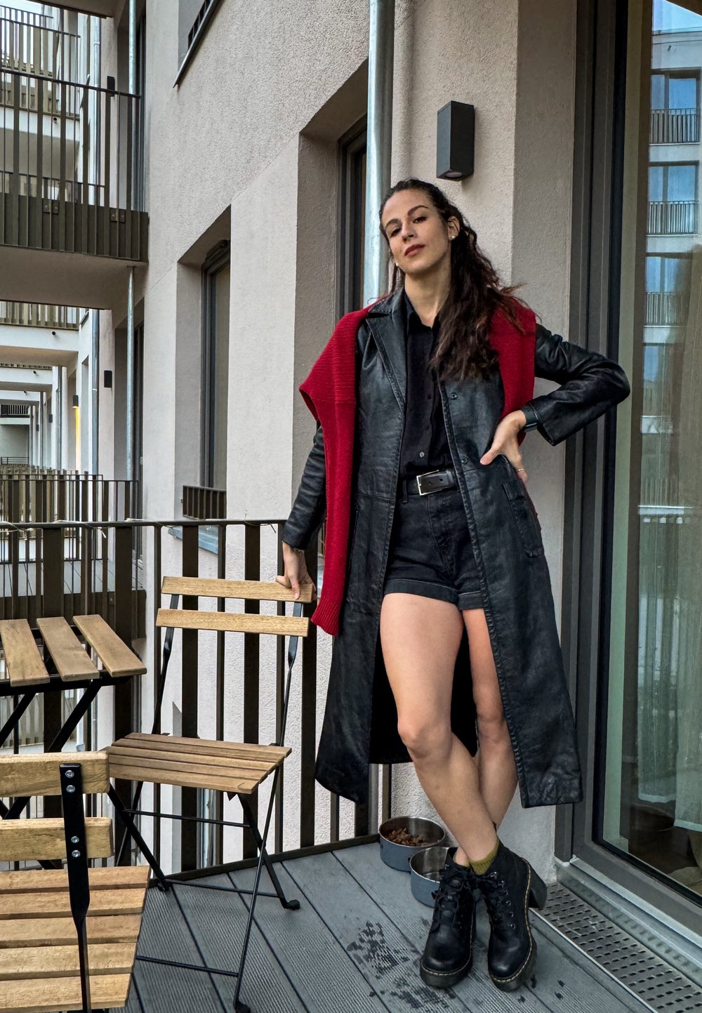 Μαύρο δερμάτινο μακρύ παλτό με κόκκινη πλεκτή λεπτομέρεια. Φοριέται πάνω από μαύρο πουκάμισο και σορτς, συνδυασμένο με μαύρες μπότες. Υπαίθρια μπαλκονάτη ρύθμιση.